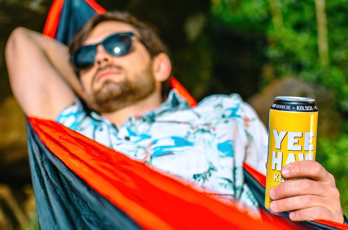 Man wearing sunglasses laying in a hammock holding a 16 ounce can of Yee-Haw Kolsch