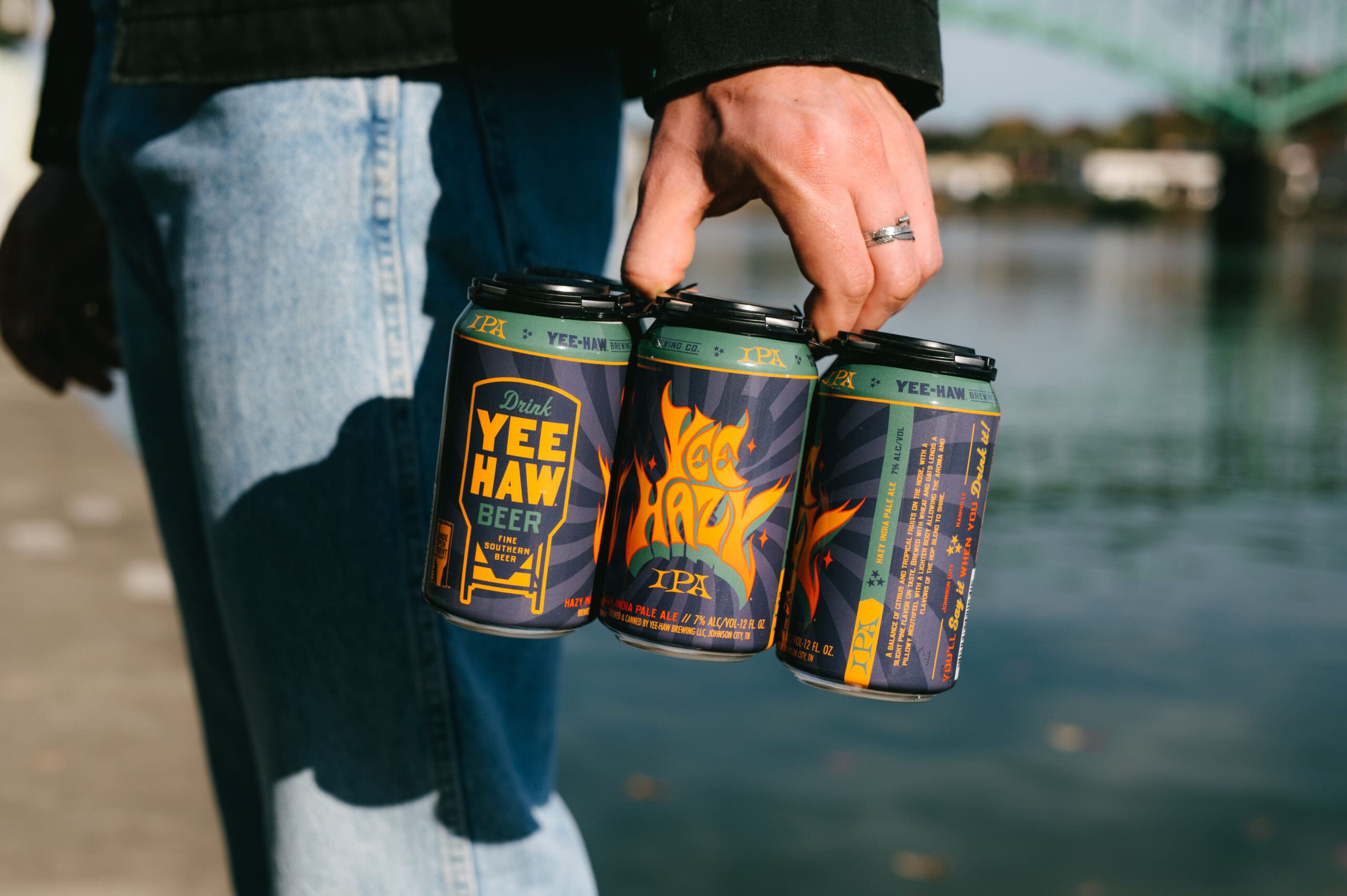 Mans hand hold a six pack of Yee-Hazy IPA by body of water